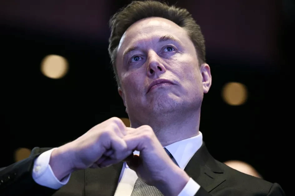 Elon Musk sur scène, micro en main, devant un écran géant lors d’une conférence, regard tourné vers le public.