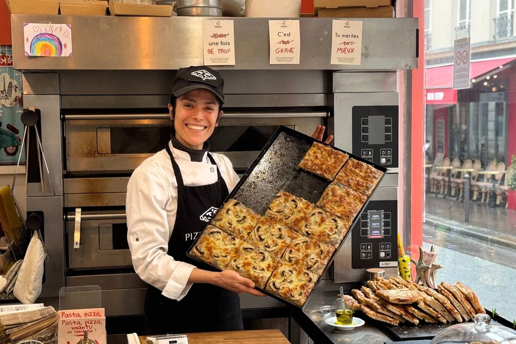 Influenceurs à Paris posant devant une pizzeria en difficulté, tenant des parts de pizza et un smartphone