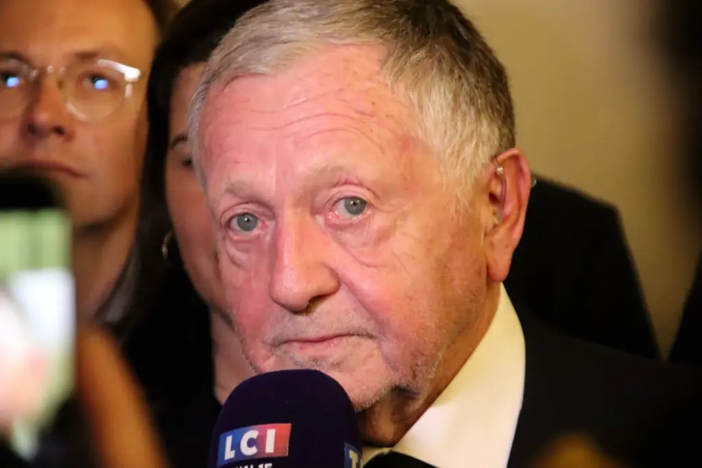 Jean-Michel Aulas en costume, micro en main, s’exprimant devant un panneau « Lyon » en arrière-plan
