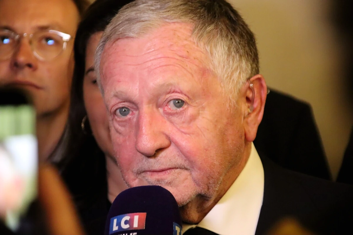 Jean-Michel Aulas en costume, micro en main, s’exprimant devant un panneau « Lyon » en arrière-plan
