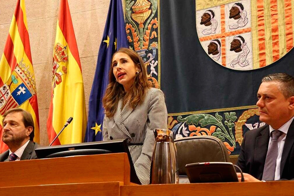 María Navarro s’exprime au pupitre des Cortes d’Aragon, devant des drapeaux et des élus assis.