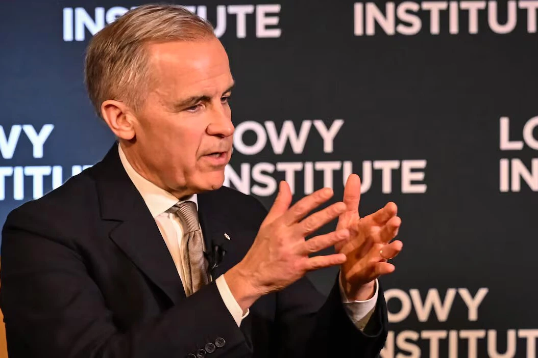 Mark Carney s’exprime devant des micros, avec Donald Trump en arrière-plan sur un écran.