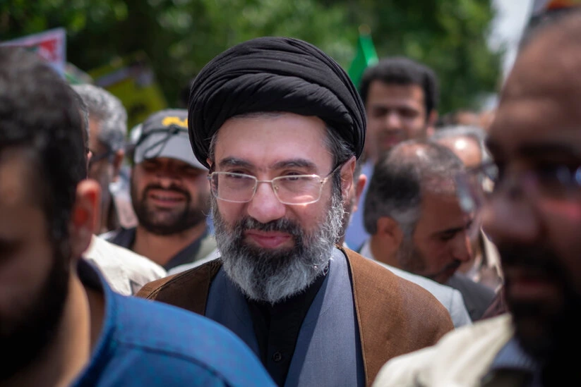 Portrait du fils d’Ali Khamenei, en tenue sombre, devant un drapeau iranien en arrière-plan