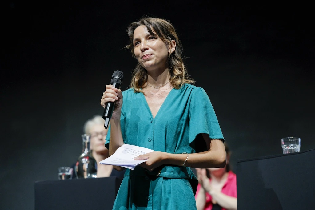 Théa Fourdrinier, candidate à Paris, posant devant un bâtiment, regard sérieux, en tenue sombre.