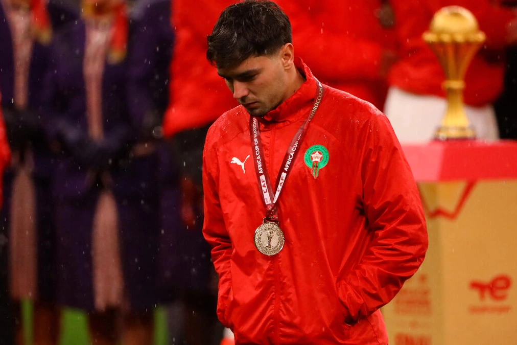 Trophée de la CAN 2025 brandi sur une pelouse, avec drapeaux du Maroc et foule en arrière-plan