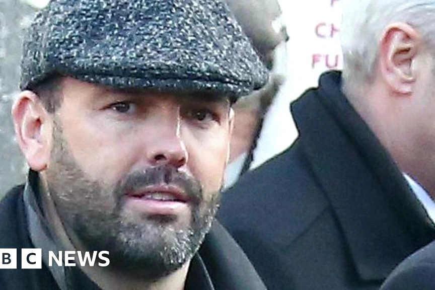 Daniel Kinahan menotté, encadré par des policiers à Dubaï, devant un bâtiment moderne éclairé la nuit