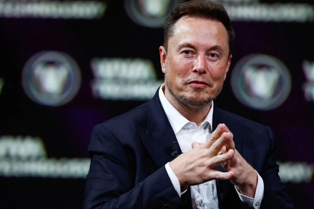 Elon Musk sur scène devant un écran Tesla, micro en main, lors d’une conférence de presse.