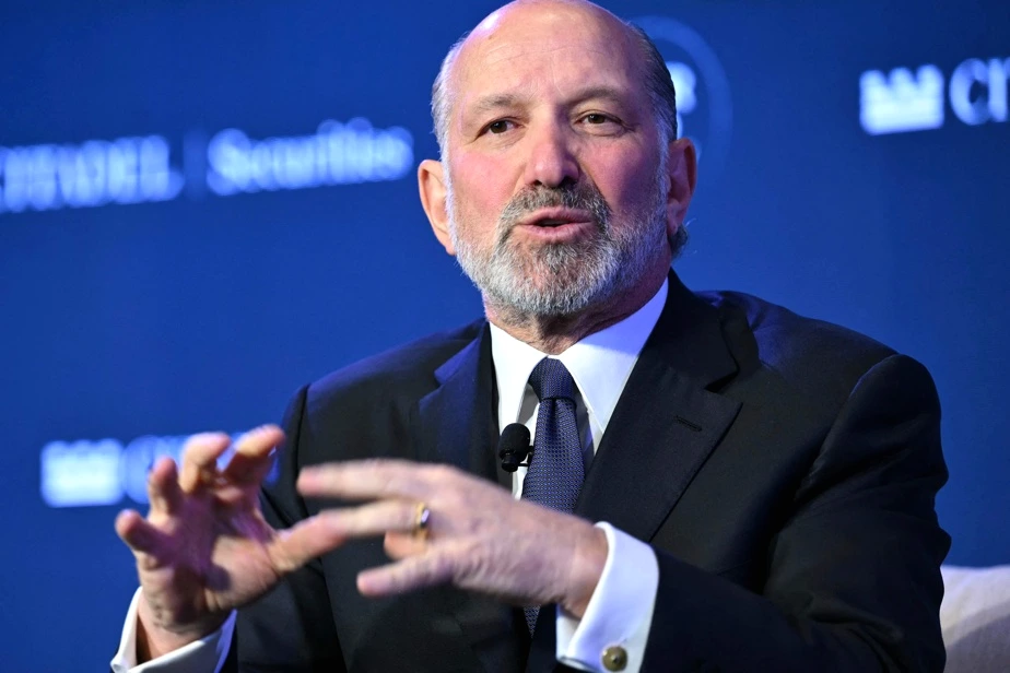 Howard Lutnick s’exprime devant des micros lors d’une conférence de presse, drapeaux en arrière-plan.