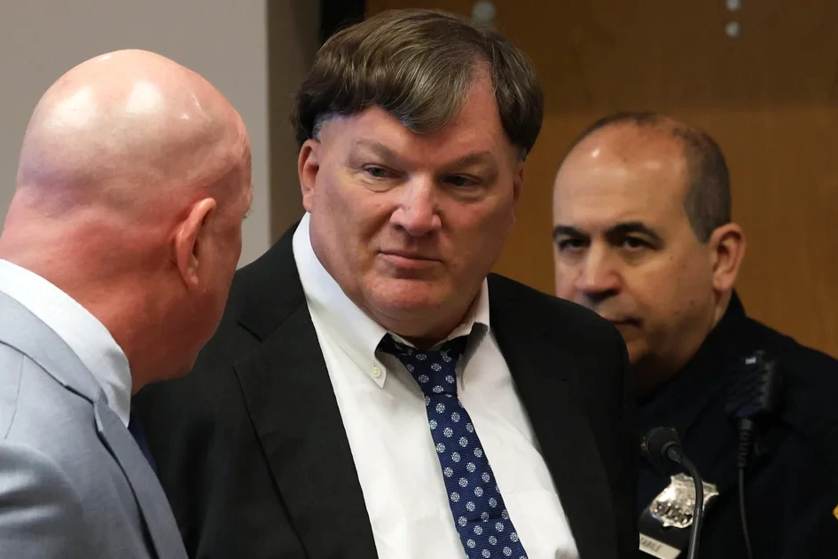 Rex Heuermann menotté, escorté par des policiers, sortant d’un tribunal sous les flashs des photographes.