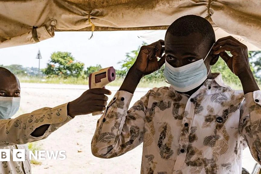 Agent de l’OMS en RDC en combinaison de protection, devant un centre de traitement Ebola et une ambulance