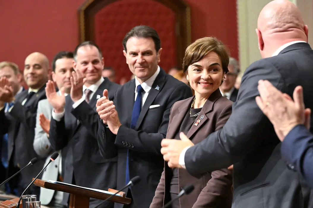 Christine Fréchette en conférence de presse, debout devant des micros, avec un drapeau du Québec en arrière-plan