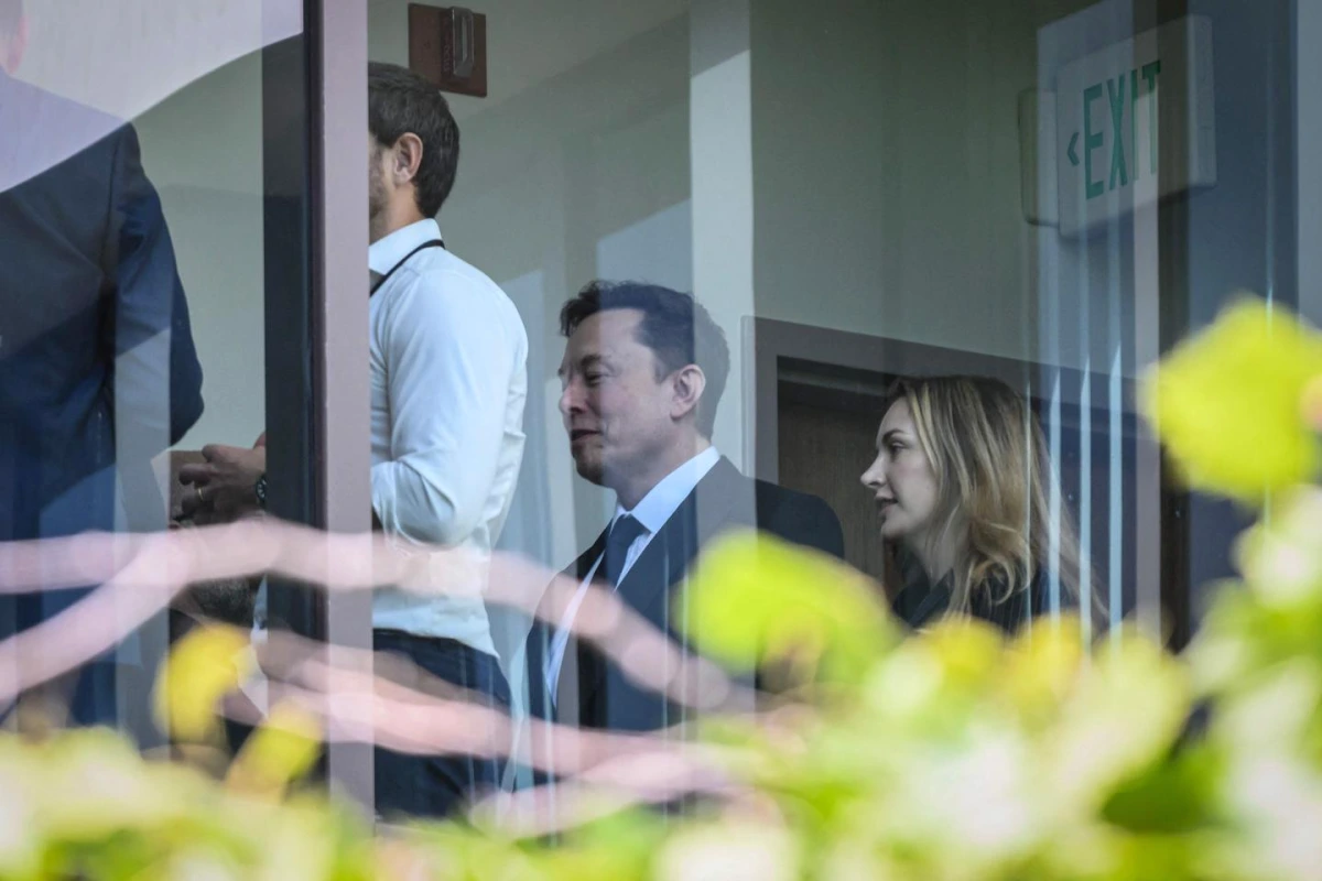 Elon Musk en costume devant un micro, avec le drapeau américain et un bâtiment officiel en arrière-plan