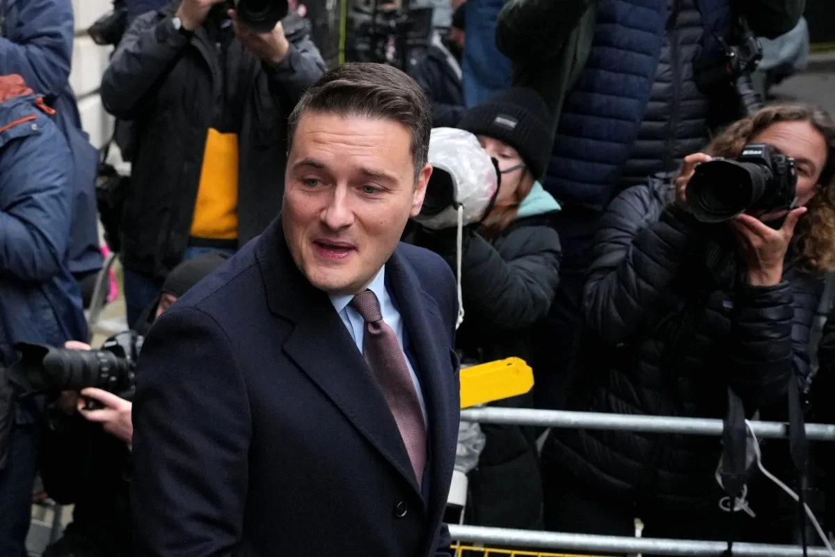 Wes Streeting en costume, parlant devant des micros, avec drapeaux britanniques en arrière-plan.