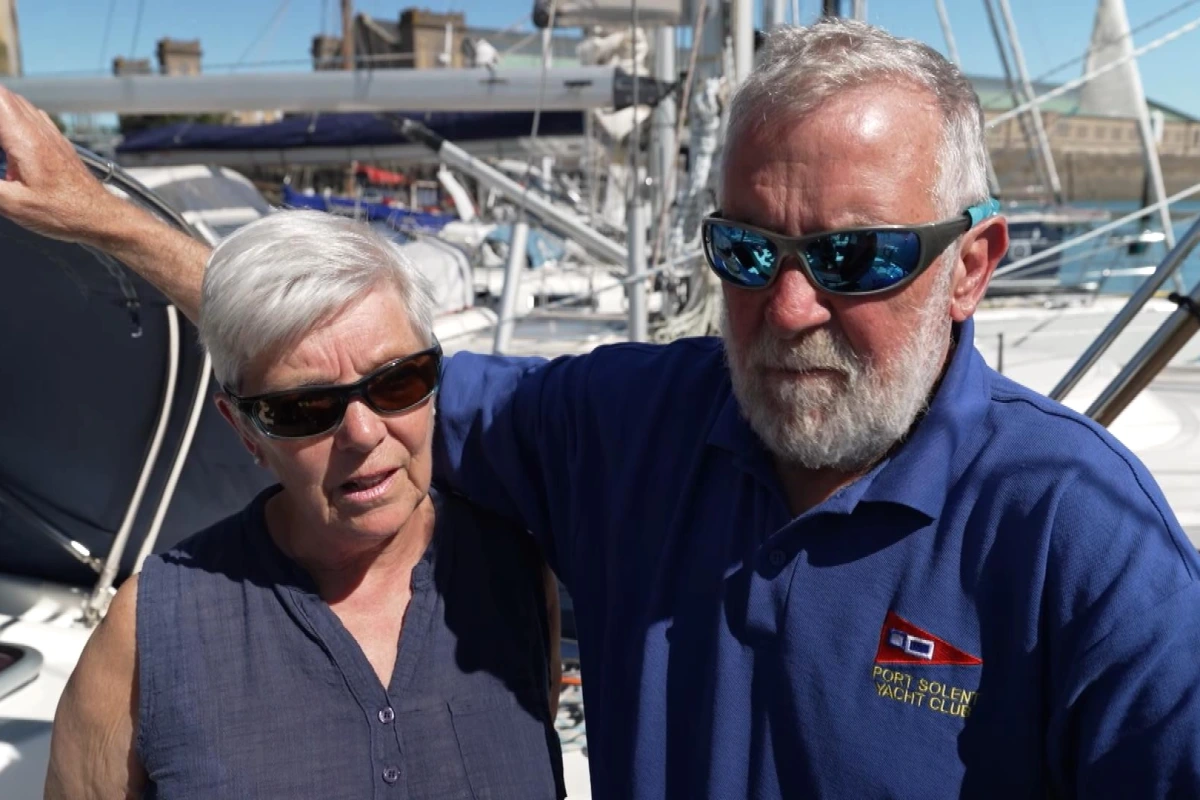 Couple britannique sur le pont d’un voilier, mer agitée, navire russe visible au loin dans la Manche