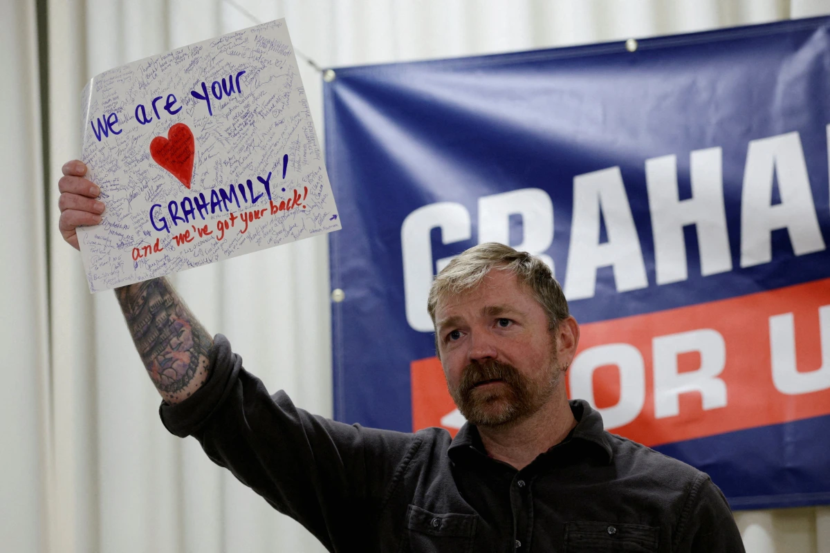 Graham Platner debout à un pupitre, micro devant lui, avec des drapeaux américains en arrière-plan
