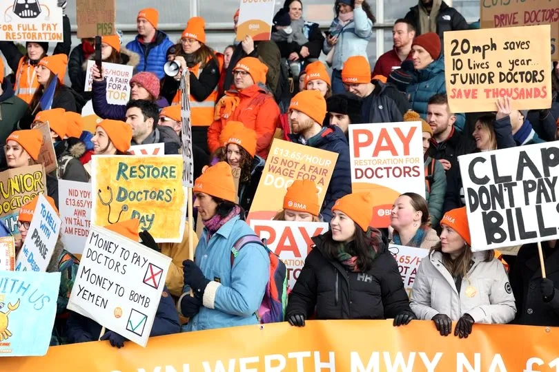 Médecins internes en Angleterre en blouses blanches, rassemblés devant un hôpital, brandissant des pancartes
