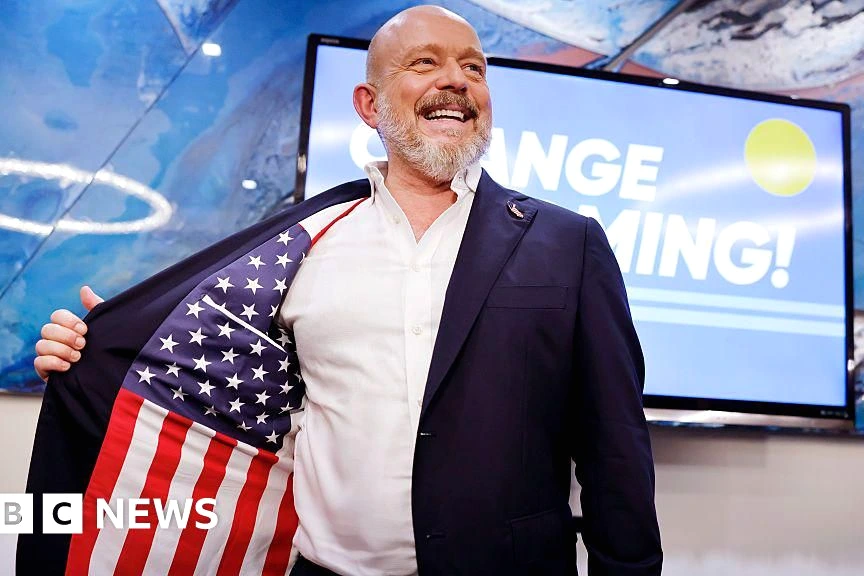 Steve Hilton en costume, debout devant un drapeau de Californie, lors d’un événement de campagne électorale